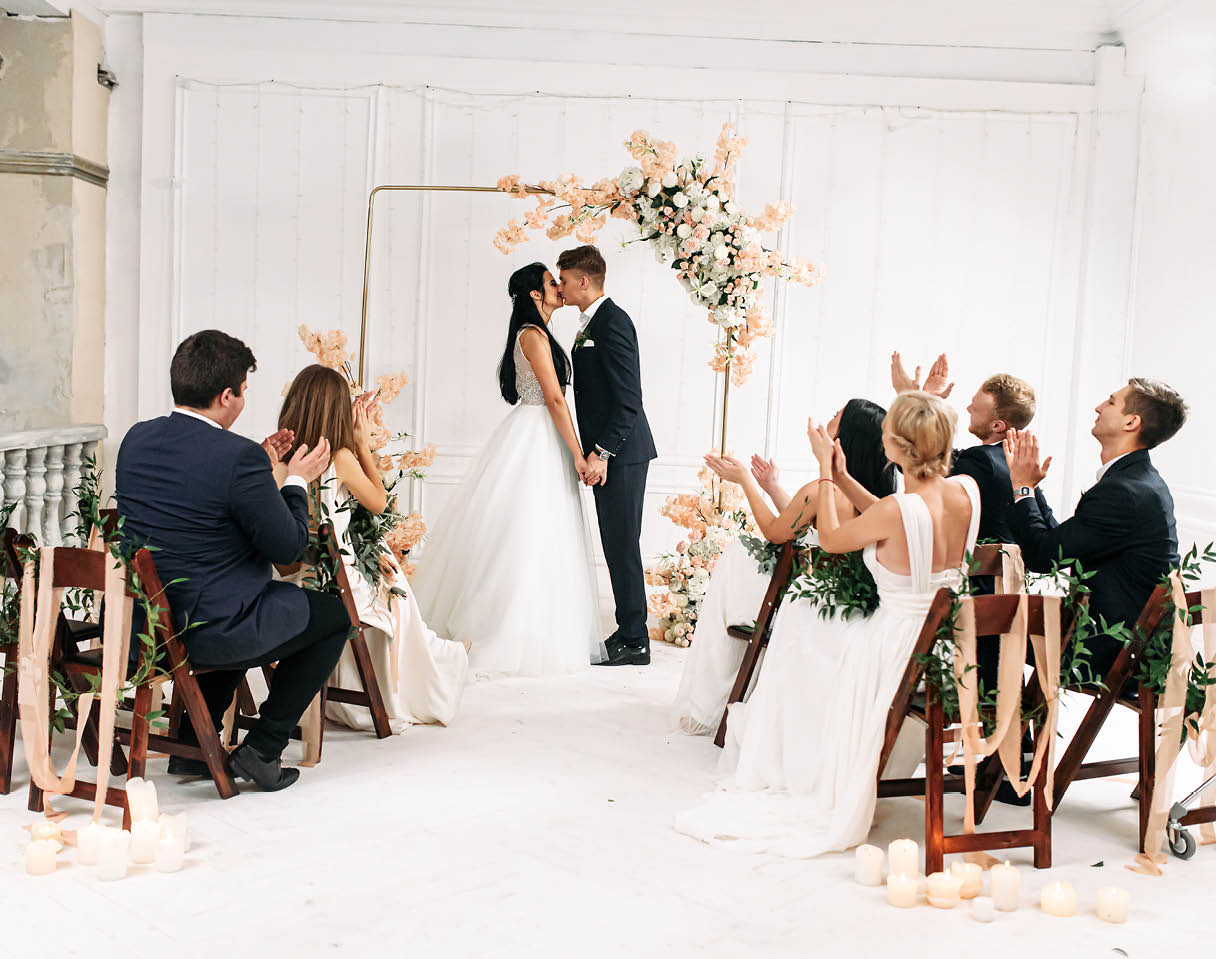 Stylish wedding ceremony decorated with flowers and green branches  Couple bride and groom near the arch hold hands and kiss, guests are sitting on chairs