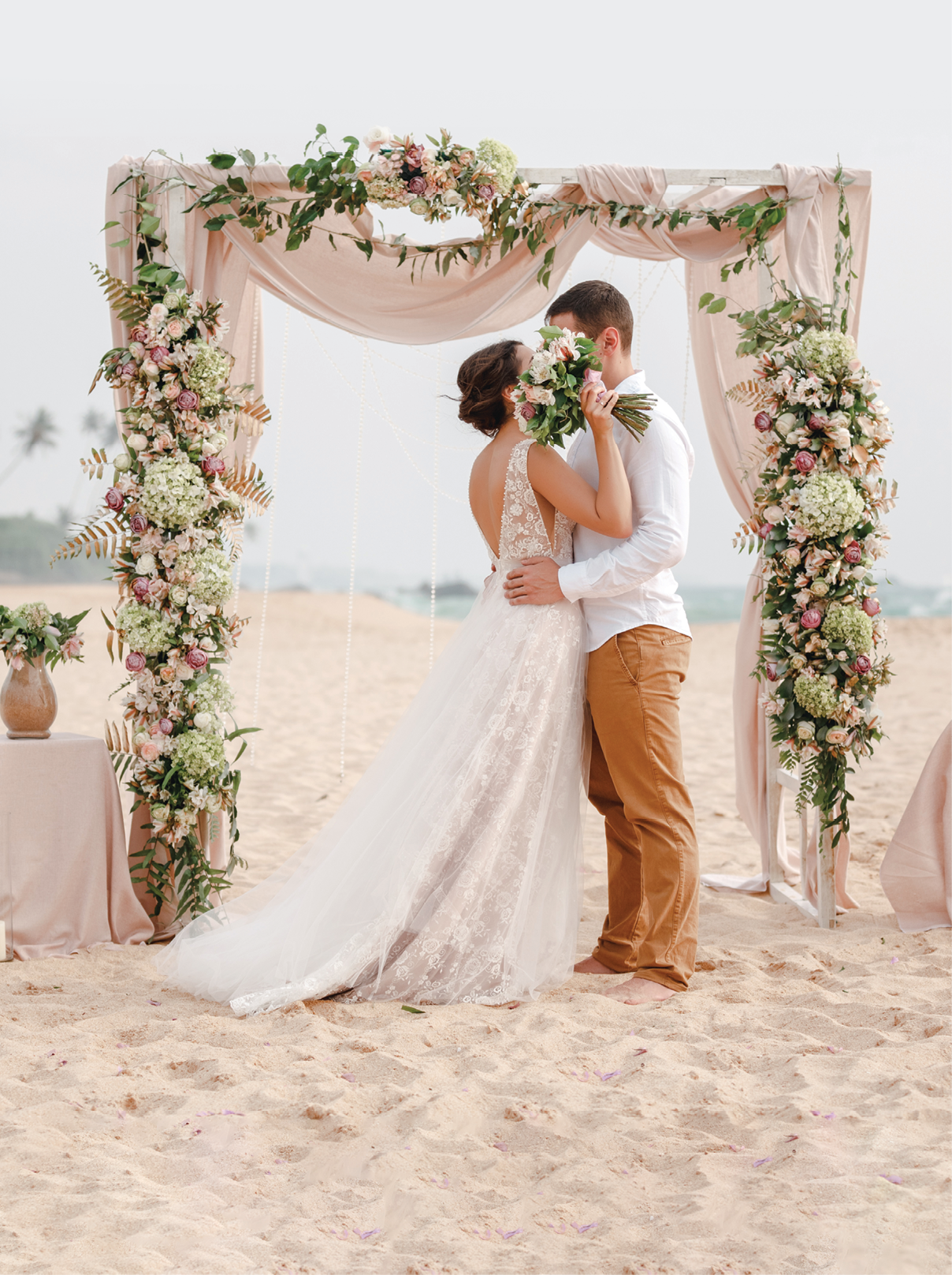 Bride and groom enjoying beach wedding in tropics, wedding arch, ocean background  Wedding ceremony on a tropical beach  Happy groom and beautiful bride kissing under the arch decorated with flowers 