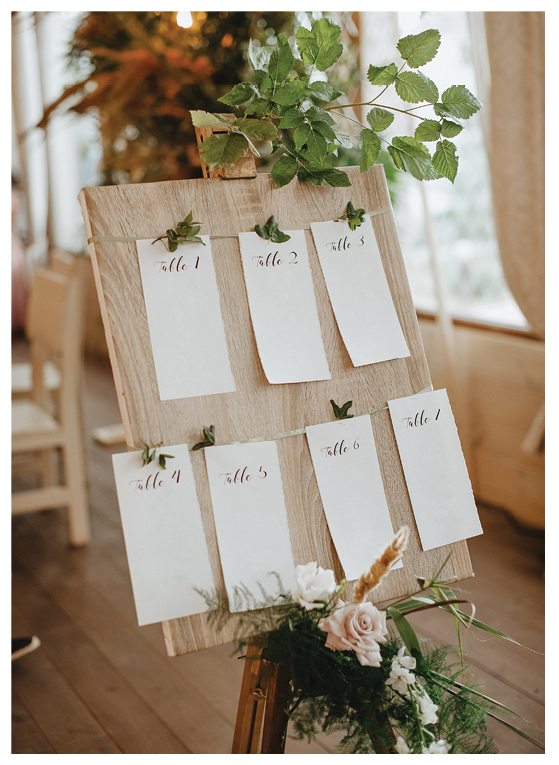 Seating plan, wooden board on an easel, paper cards with table numbers, a composition of flowers and tropical leaves, raspberry branches.