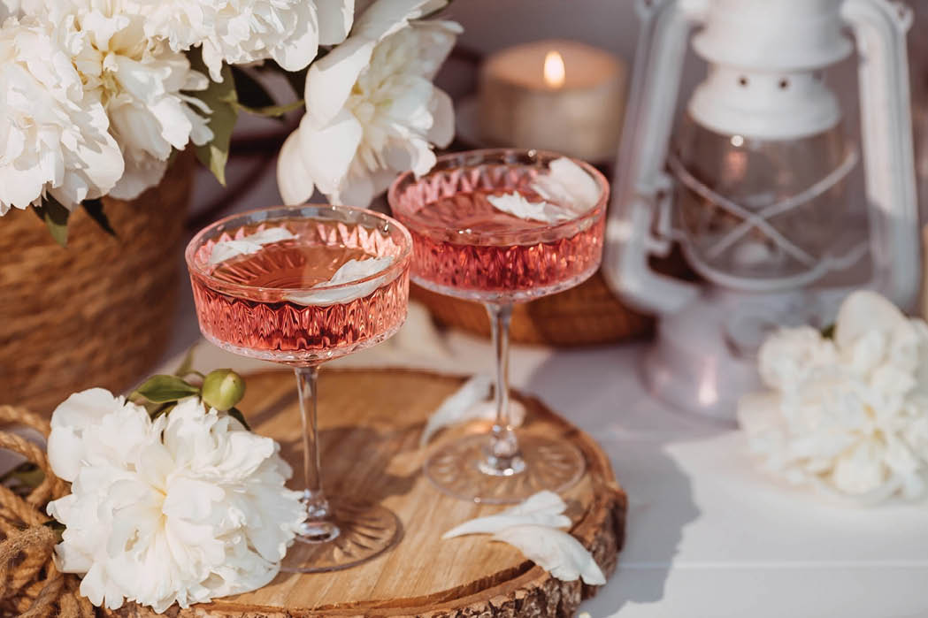 A bouquet of white peonies, flower petals, candles and a fashionable glass for dessert and champagne with a pink drink inside. The concept of a party and a holiday.