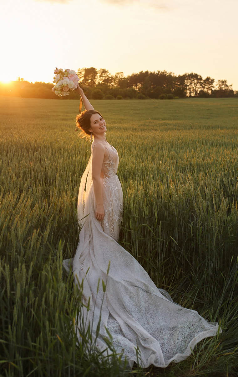 Young happy bride in lace wedding dress holding bouquet of flowers in her hand and posing in the field in the summer sunset