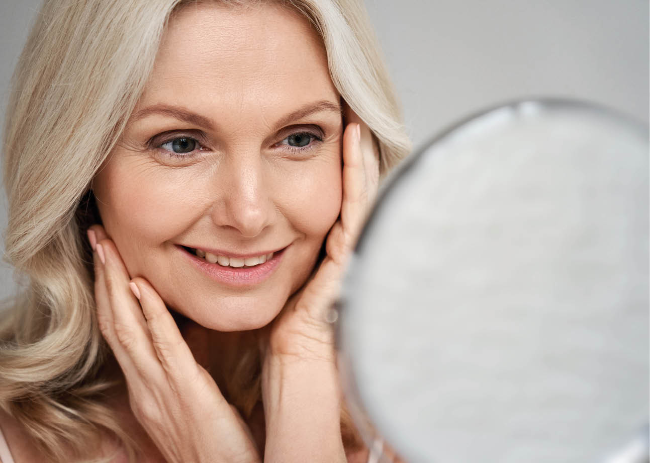 Happy 50s middle aged woman model touching face skin looking in mirror  Smiling mature older lady pampering, enjoying healthy skin care, aging beauty, skincare treatment cosmetic products concept 