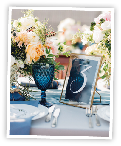 Long dinner tables covered with white cloth, served with porcelain and blue glasses and rich decorated with flowers stand on the roof of a house
