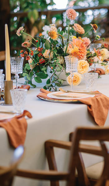 A large, long, decorated, wooden table and chairs, covered with a white tablecloth with dishes, flowers, candles, stands outdoor near the forest in nature. Wedding banquet. lights on background