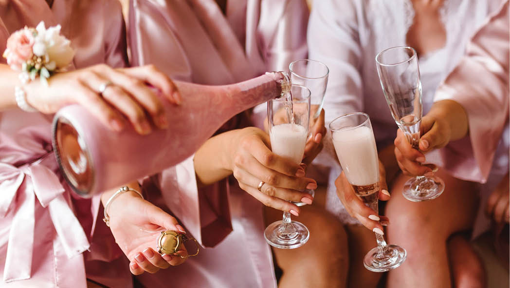 Young bridesmaids clinking with glasses of champagne in hotel room. Closeup photo of cheerful girls celebrating a bachelorette party. Females have toast with wine.