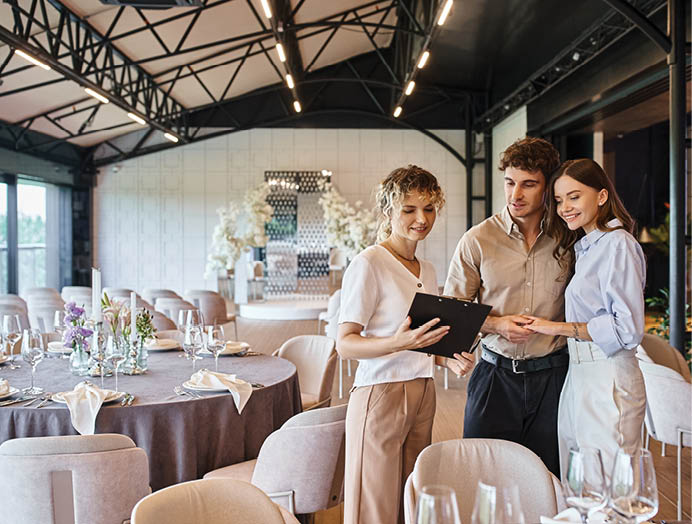 happy couple looking at contract on clipboard near banquet coordinator in decorated event hall