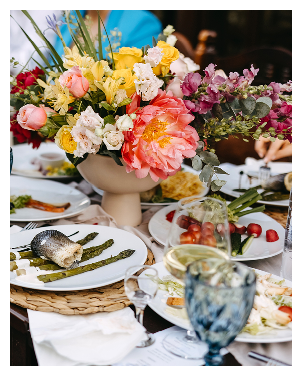 Festive table setting with colorful floral centerpiece, guests dining, fish dish, asparagus, wine, water, and vibrant atmosphere.