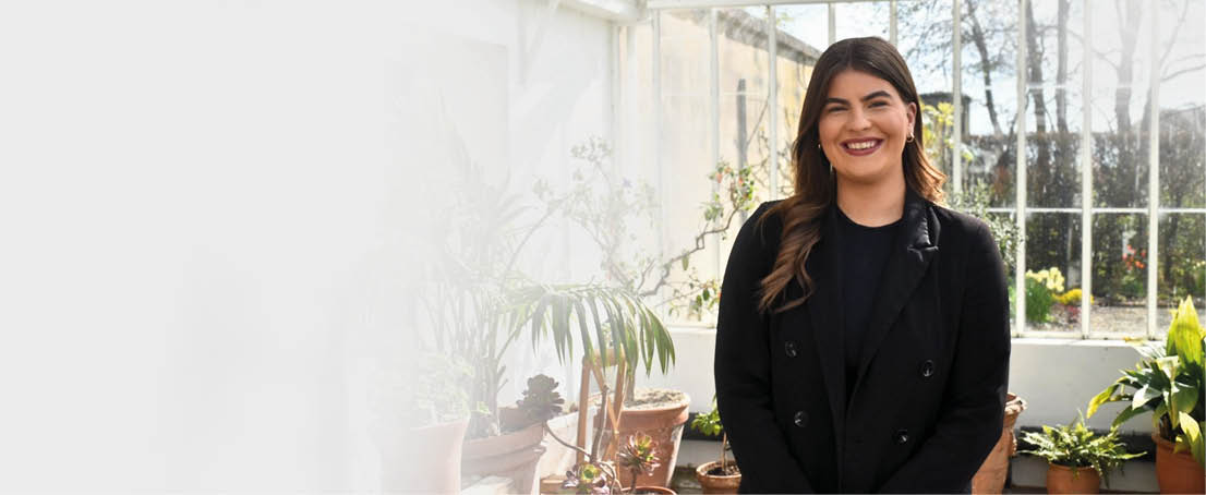 A woman in a black jacket is standing in front of a row of potted plants. Description generated by AI