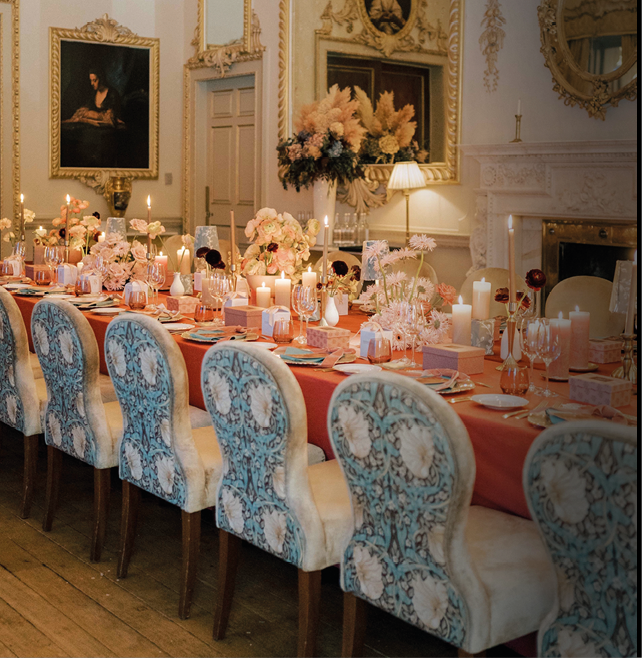 A long dining table with a coral tablecloth is elegantly set with floral arrangements, candles, glassware, and patterned upholstered chairs in a richly decorated room with framed paintings and ornate mirrors. Description generated by AI