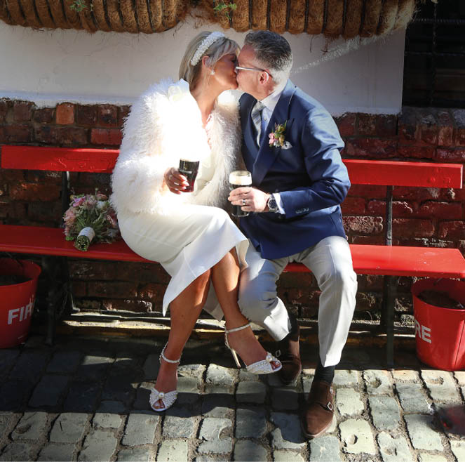 Two adults dressed in formal attire are sitting on a red bench outdoors, sharing a kiss while holding glasses of dark beer. Description generated by AI