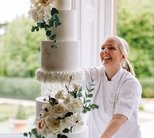 A woman is smiling while standing in front of a three-tiered cake. Description generated by AI