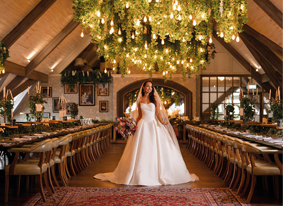 A person in a white wedding dress holding a bouquet stands in the center of a warmly lit, elegantly decorated banquet hall with long tables and hanging greenery and lights. Description generated by AI