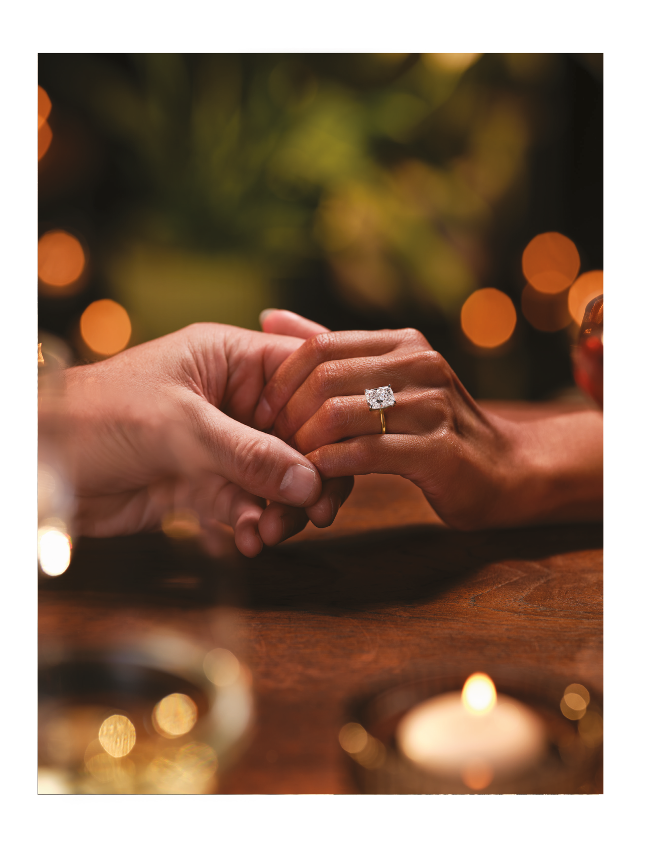 A close-up of two people holding hands across a table with a lit candle and a blurred background, alongside an elegant gold ring with a large rectangular diamond and the text “Fill life with love." Description generated by AI