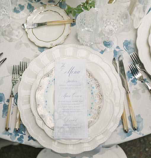 A table with a blue and white tablecloth, a plate with a napkin, and a vase of flowers. Description generated by AI