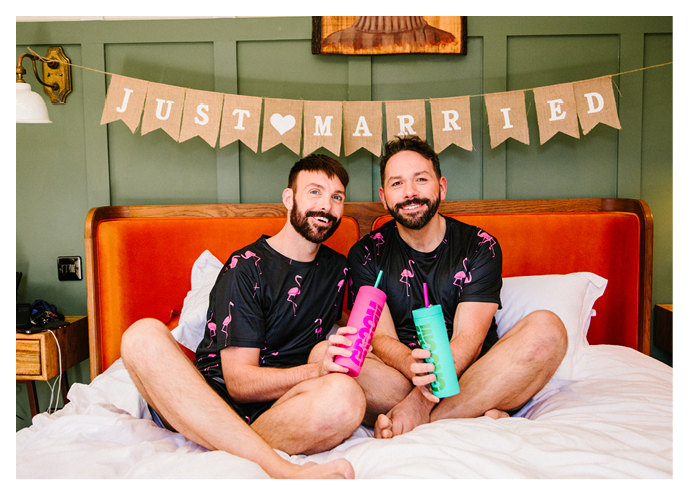Two men sitting on a bed with a banner that says “Just married". Description generated by AI
