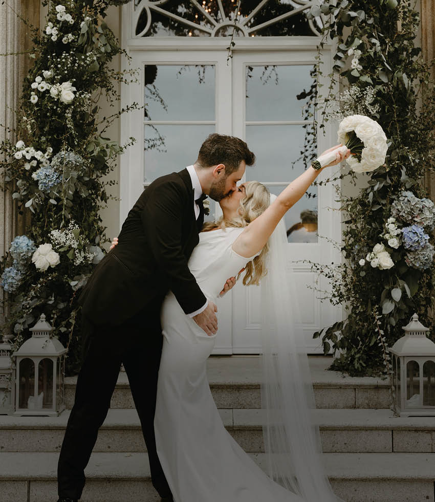 A bride and groom are kissing in front of a doorway with a flower arrangement. Description generated by AI
