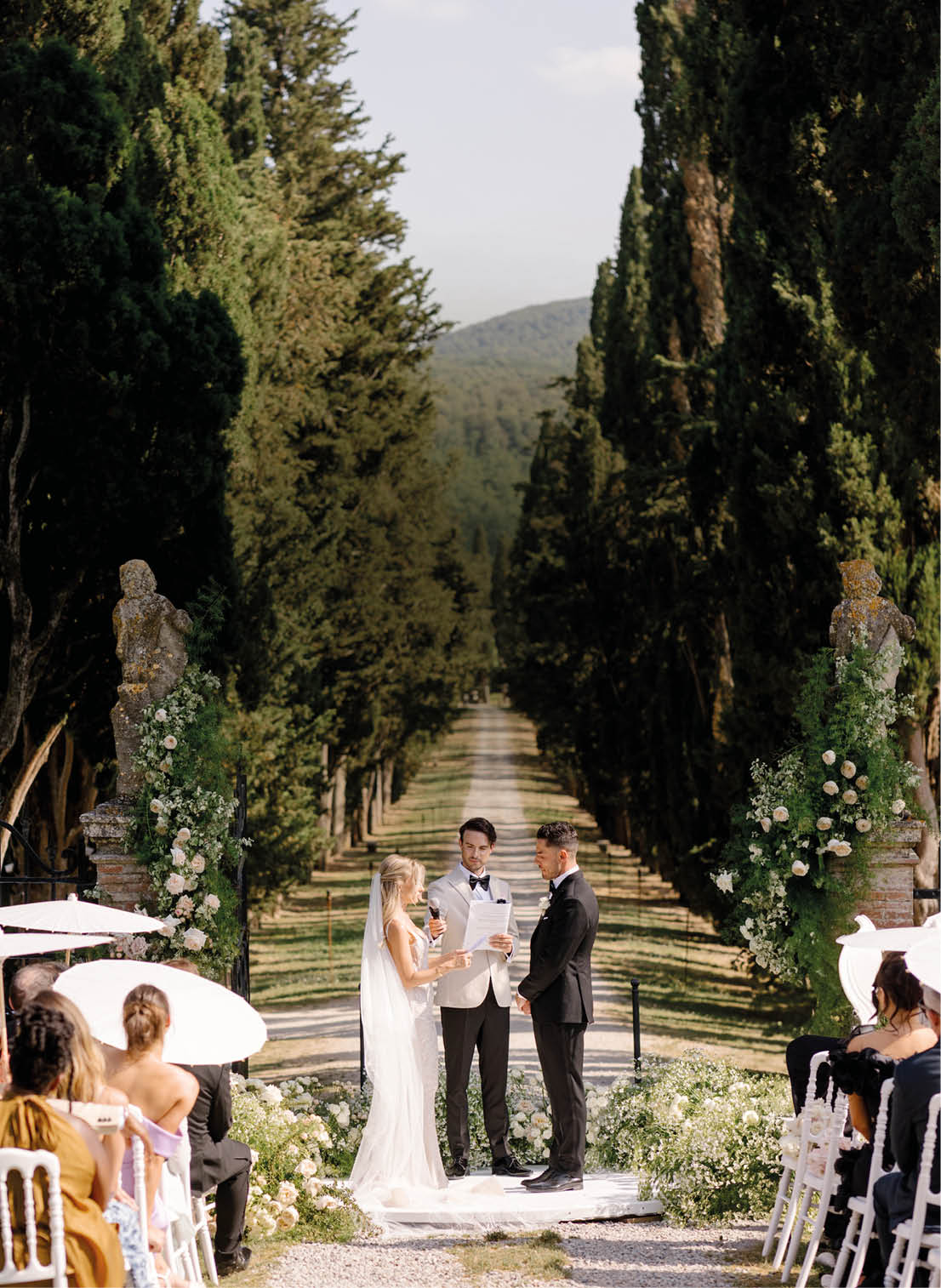 A bride and groom stand in front of a row of trees. Description generated by AI