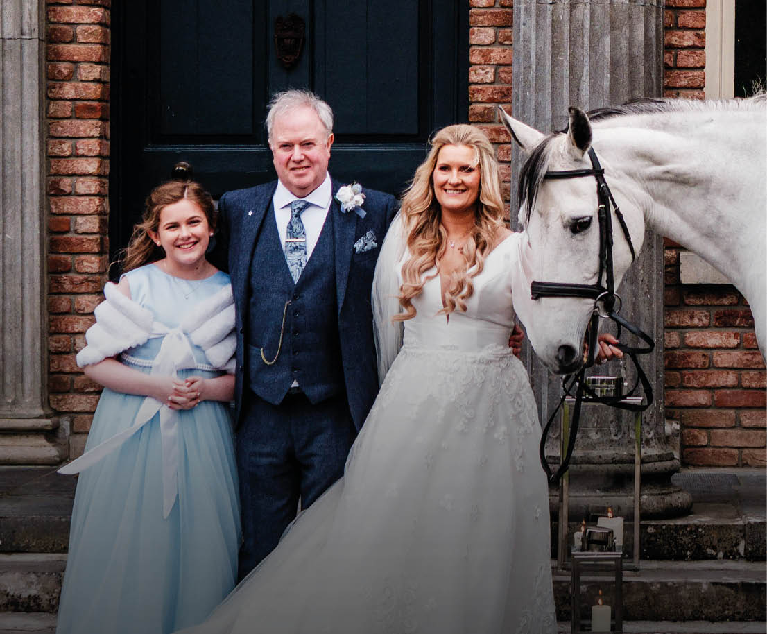 A bride and groom pose with a white horse in front of a brick building. Description generated by AI