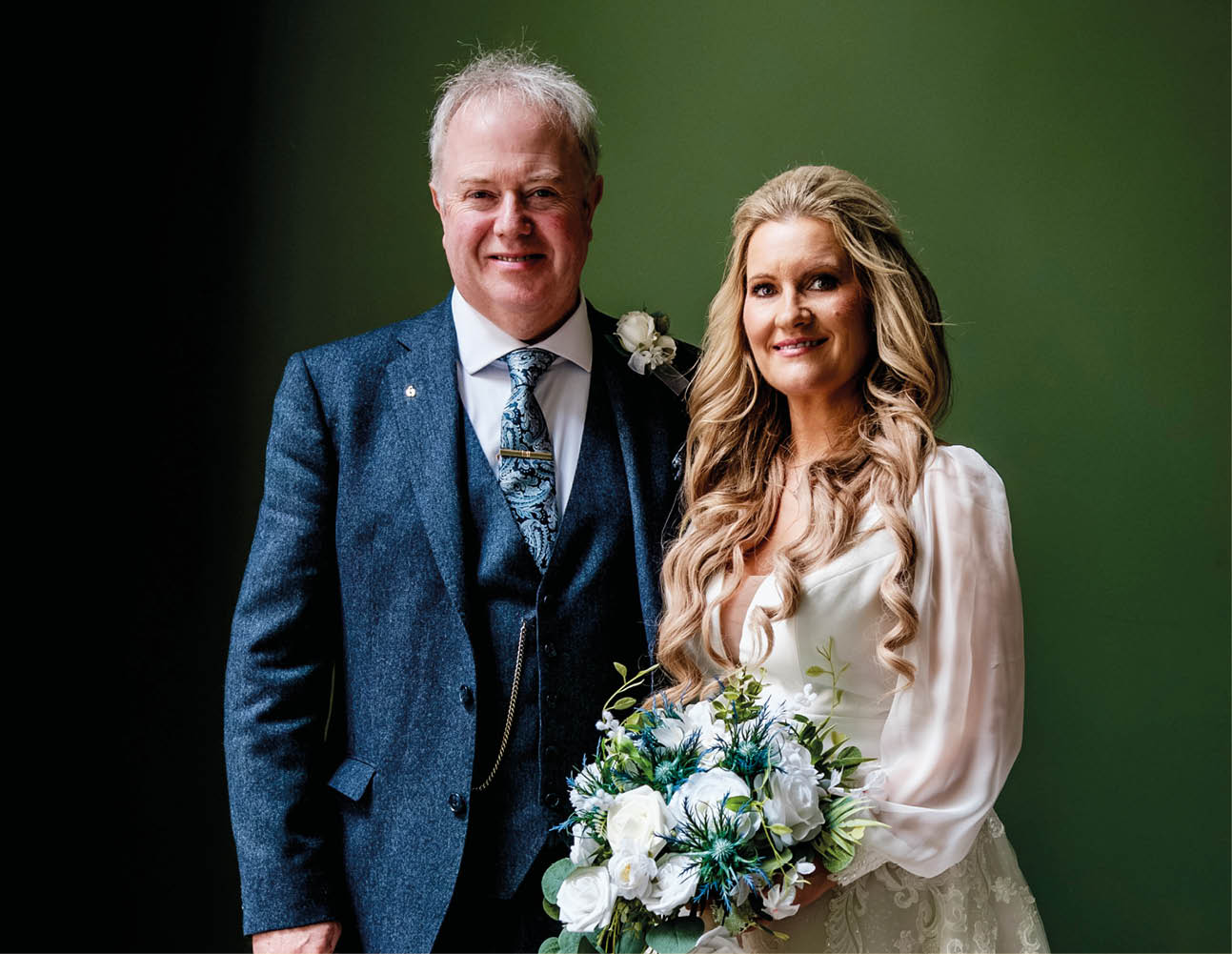 A bride and groom are standing together in front of a green wall. Description generated by AI