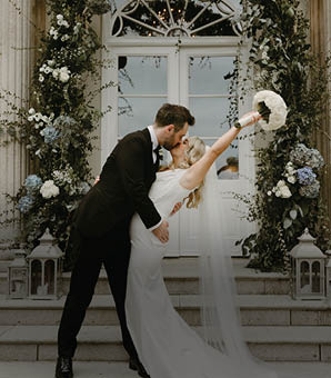 A bride and groom standing in front of a building. Description generated by AI
