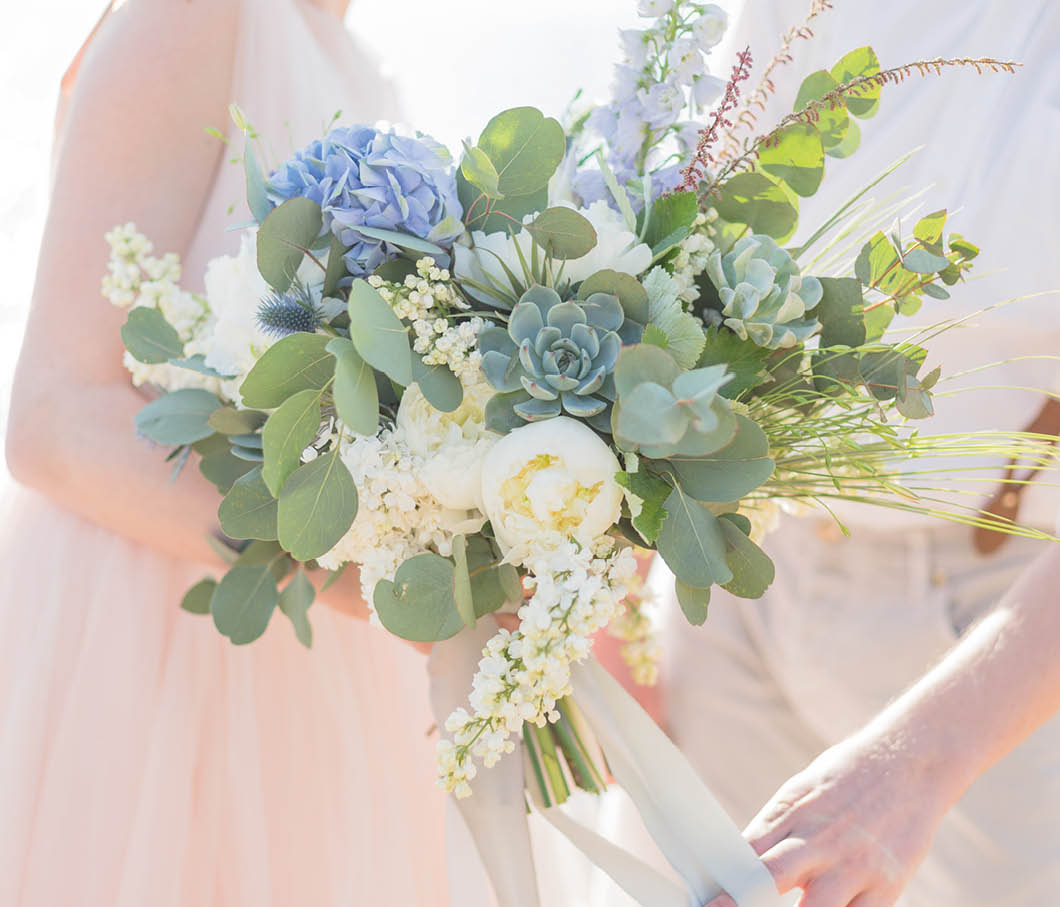 A bride and groom are holding hands and posing for a picture. The bride is wearing a white dress and the groom is wearing a white shirt. The bride is holding a bouquet of flowers. Description generated by AI