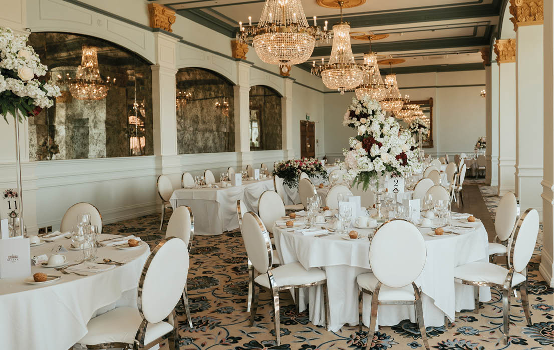 A large room with a chandelier and a long table with white tablecloths and place settings. Description generated by AI