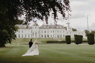A bride and groom are standing in a field with a large white house in the background. Description generated by AI