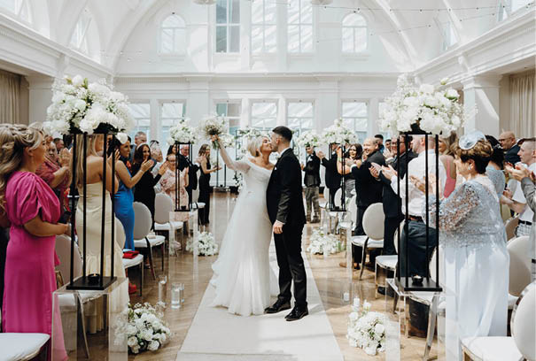 A bride and groom stand in front of a crowd of people. Description generated by AI