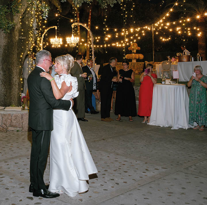 A bride and groom are dancing under a tree with a crowd of people watching. Description generated by AI