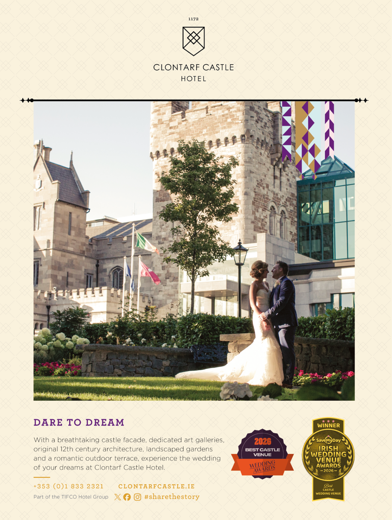 Two adults dressed in formal wedding attire stand closely together holding hands in a garden area in front of a historic castle building with flags and greenery. Description generated by AI