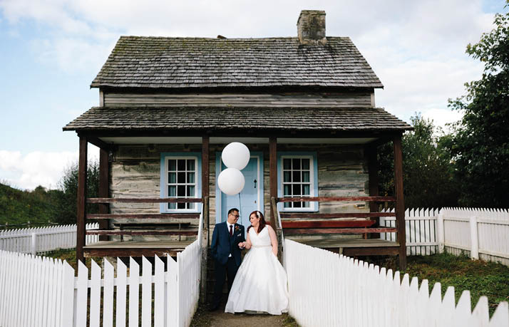 A bride and groom standing in front of a white fence. Description generated by AI
