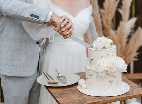 Bride and groom cutting stylish wedding cake at wedding in outdoor. Wedding couple holding knife and cutting together wedding cake decorated with flowers