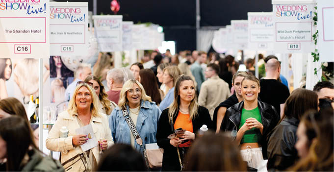 A crowded indoor event with many people walking and standing among booths displaying signs for a wedding show. Description generated by AI