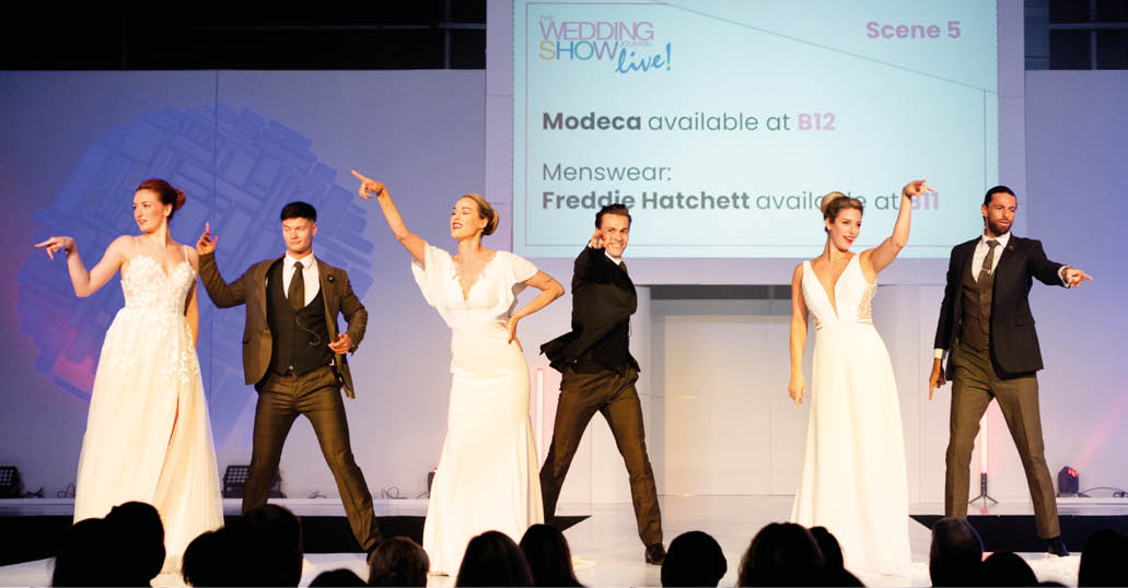 Five adults dressed in formal attire stand on a stage with a screen behind them displaying text about a wedding show and fashion brands. Description generated by AI