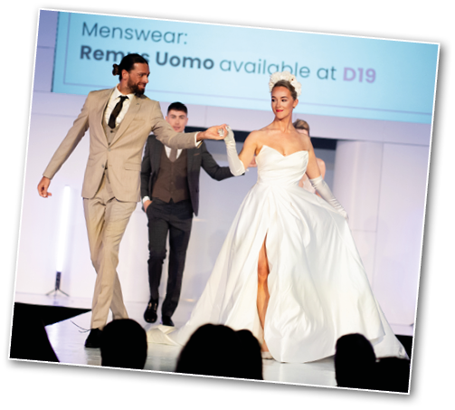 Three adults are walking on a runway with one wearing a white formal gown with a high slit and the others dressed in suits. Description generated by AI