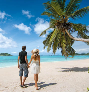 Beautiful young couple relaxing on an exotic beach from Seychelles. Couple in love travel on honeymoon on exotic island. Young lovers under palm tree and surrounded by blue sea from Seychelles Islands