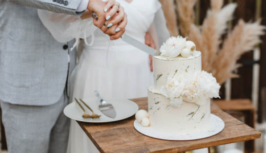 Bride and groom cutting stylish wedding cake at wedding in outdoor. Wedding couple holding knife and cutting together wedding cake decorated with flowers