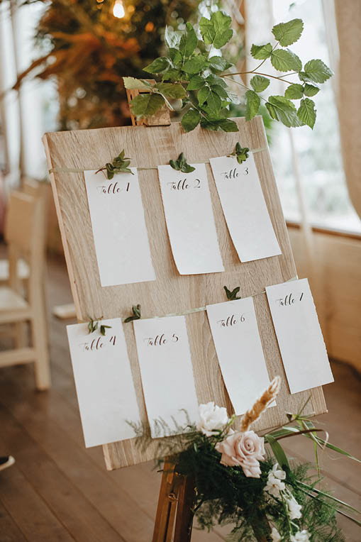 Seating plan, wooden board on an easel, paper cards with table numbers, a composition of flowers and tropical leaves, raspberry branches.