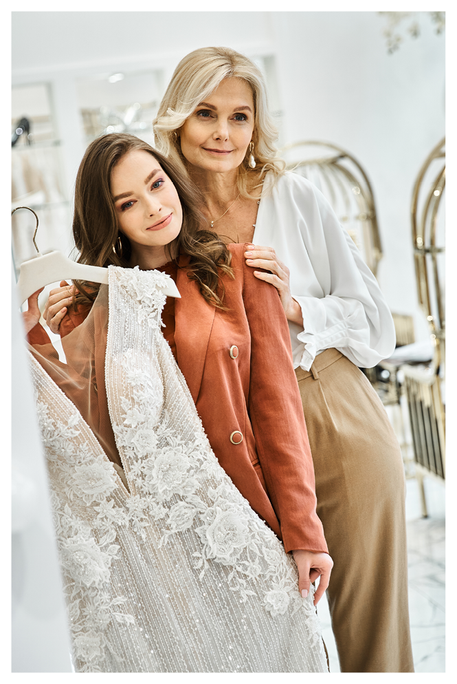 Two women, a young bride and her mother, stand next to each other in front of a mirror, browsing wedding attire.