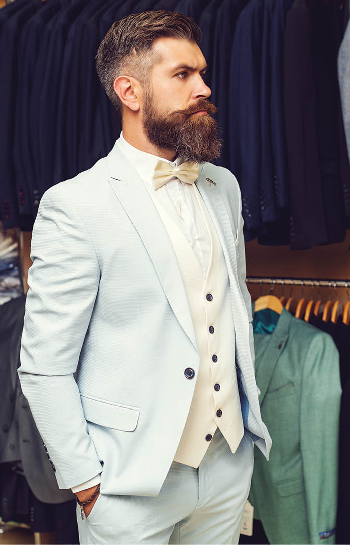 Sale, shopping. Handsome bearded brutal man in classical blue costume suit. Male suits hanging in row. Men clothing, boutiques. Tailor, tailoring. Stylish men's suit. Man suit, tailor in his workshop