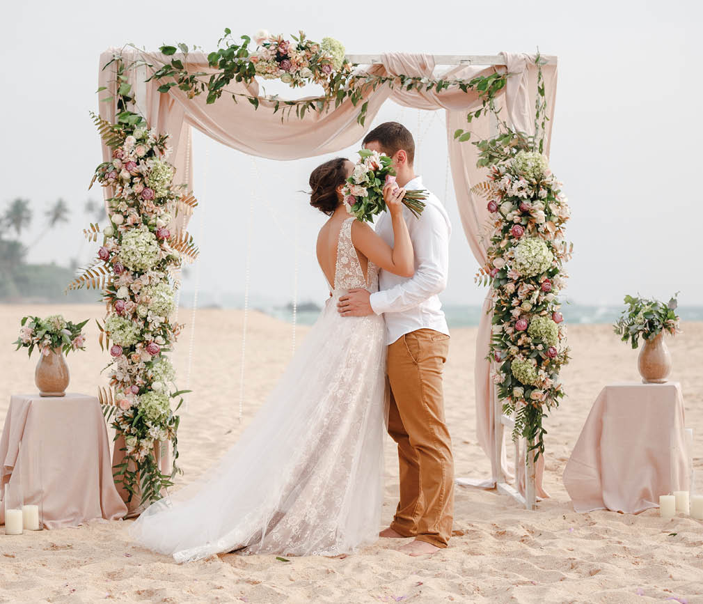 Bride and groom enjoying beach wedding in tropics, wedding arch, ocean background. Wedding ceremony on a tropical beach. Happy groom and beautiful bride kissing under the arch decorated with flowers 