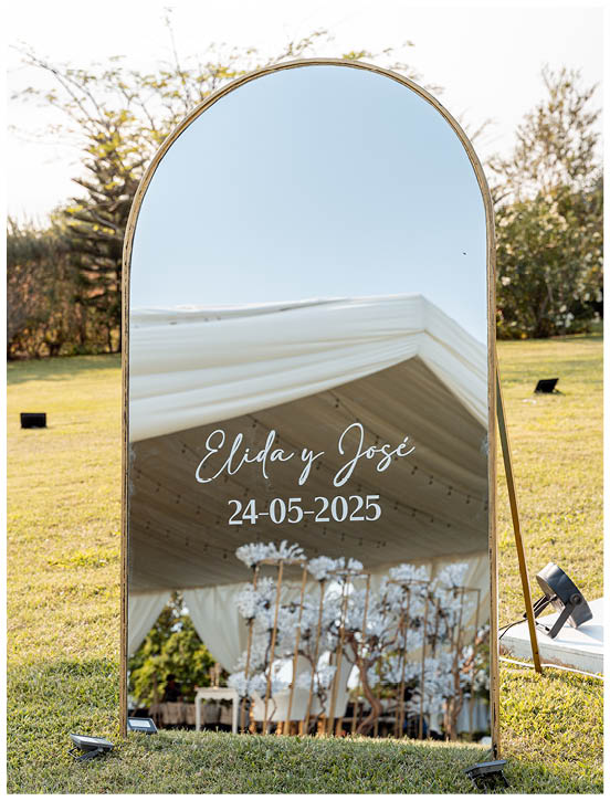 Beautiful personalized mirror with gold borders, displaying the couple's names, located in an event venue garden. Wedding decor, green grass, and plants are visible. 
