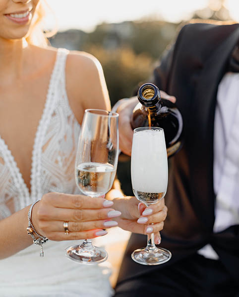 Close-up photo of newlyweds hands, men and women hold glasses of champagne or white wine. Drink is poured. Wedding day romantic evening on rooftop against background of city. Love