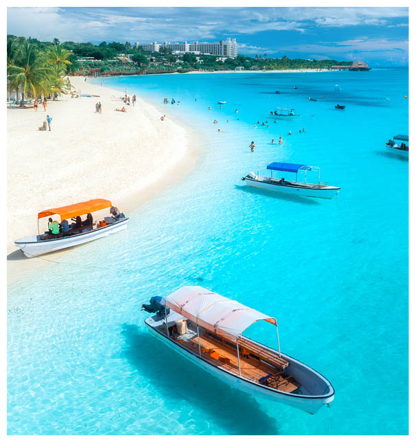 Aerial view of the boats on tropical sea coast with white sandy beach on summer sunny day. Vacation in Kendwa, Zanzibar. Landscape with boat, yacht, clear water, green palms, blue sky. Top drone view