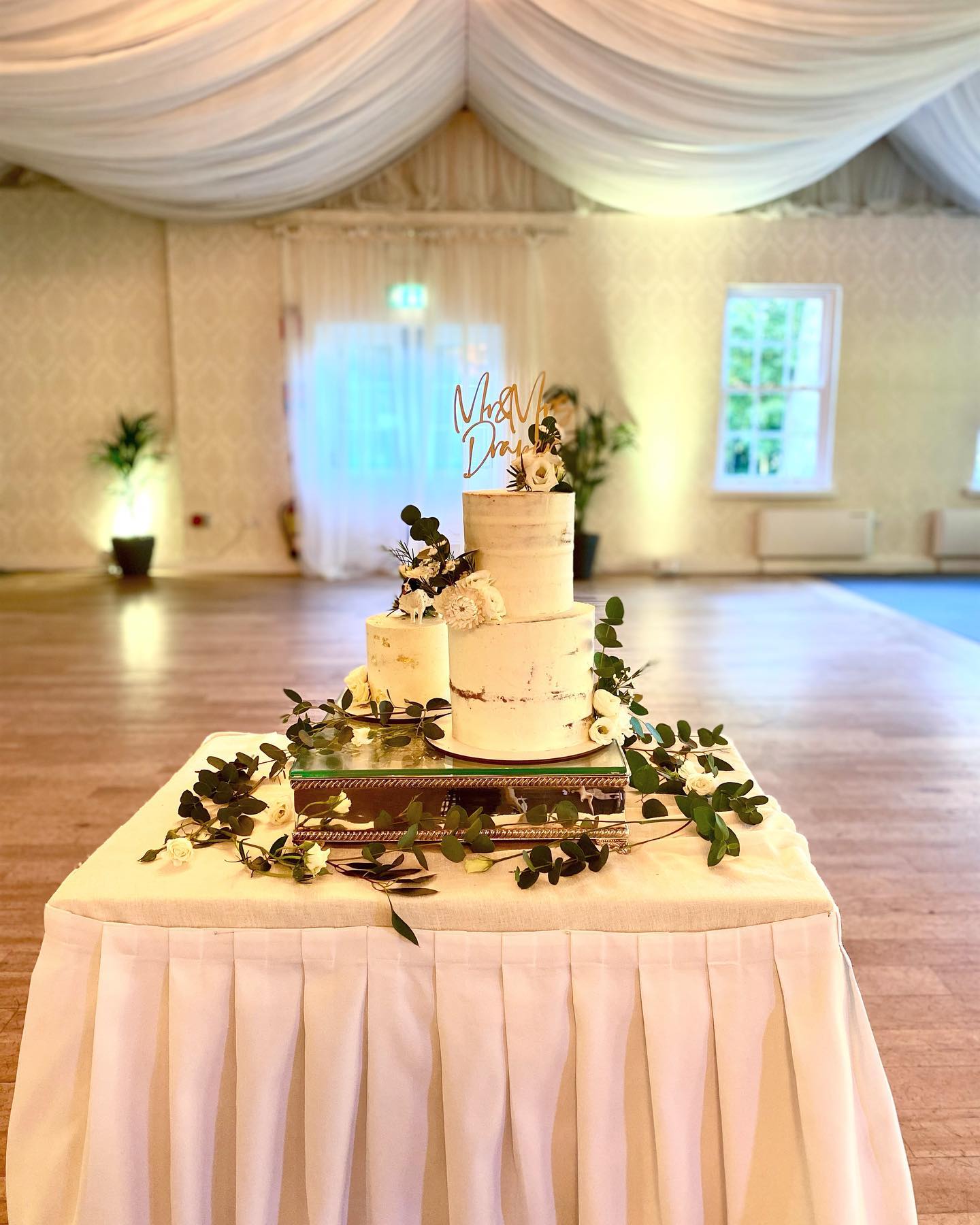 This image highlights the wedding trends of 2022 that we are predicting. This is of a semi-naked cake. Green leaves surround the cake with a gold cake topper on the final tier.