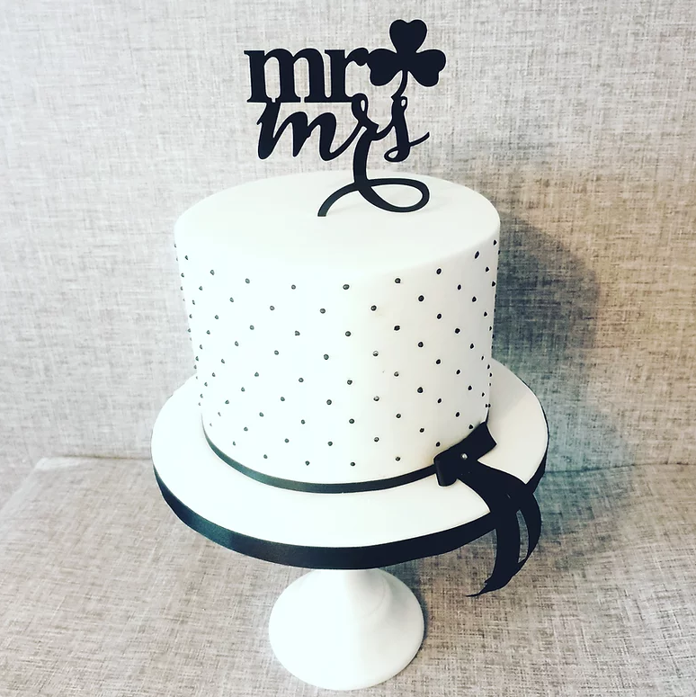 This is a monochrome single tier cake. A white cake with black decorative dots sits on a cake stand with a black bow around the bottom of the cake and a black topper on the top. Image used in the 6 Wedding Cake Trends of 2022 article.