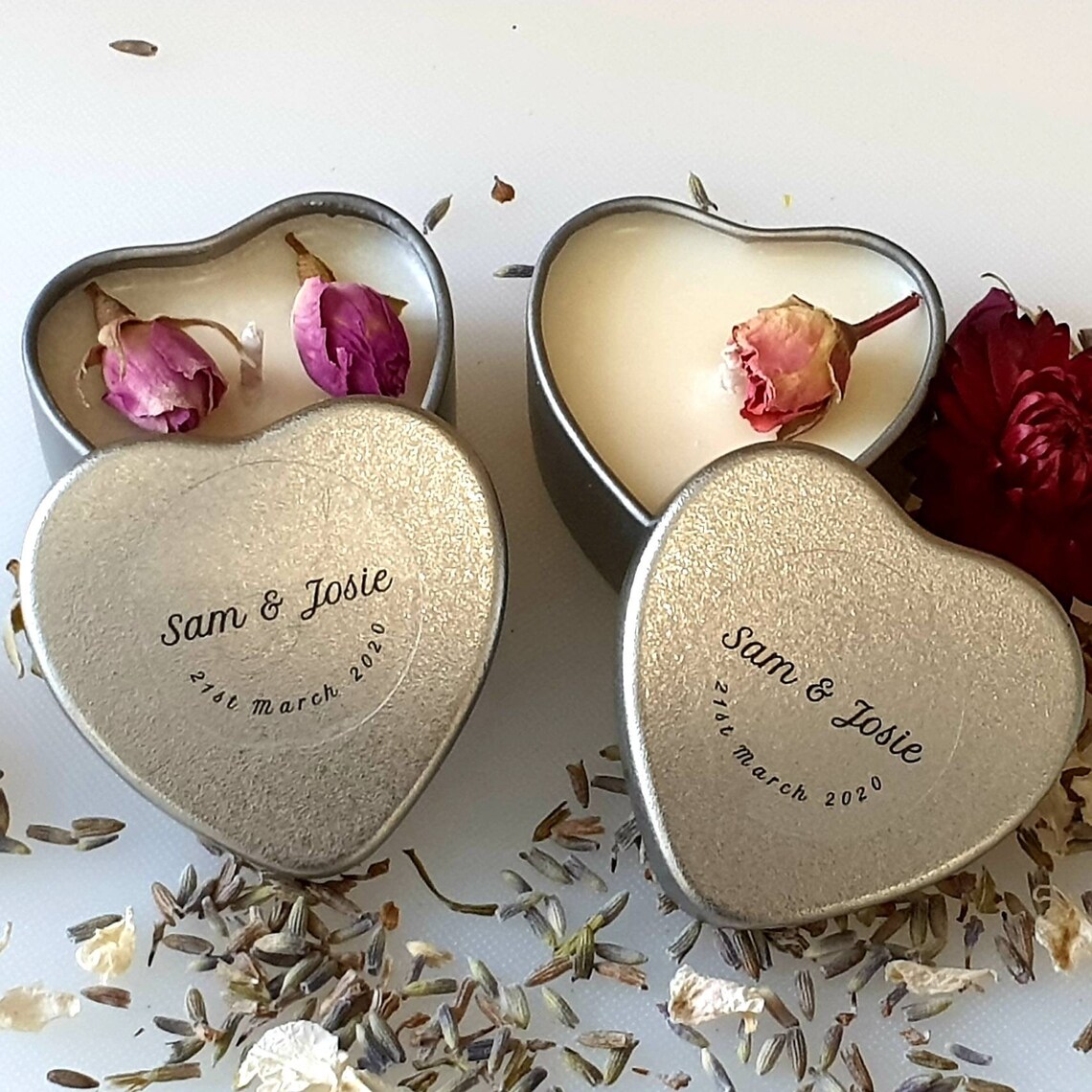 Two small personalised candles sit side by side. The top of the candle states the name of the couple and the date they got married. Flower confetti is seen inside the candle. Image used in the Cute Wedding Favours Your Guests Will Want To Take Home article. 