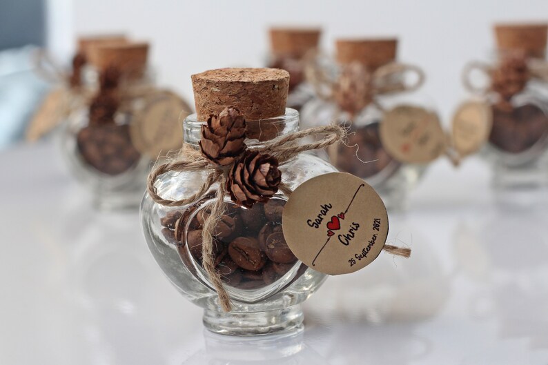 This image shows a small jar of coffee beans. A small piece of rustic rope is tied around the top of the jar with two mini acorns. A cardboard cutout is personalised and highlights the name of the couple and the date of their wedding. Image used in the Cute Wedding Favours Your Guests Will Want To Take Home article