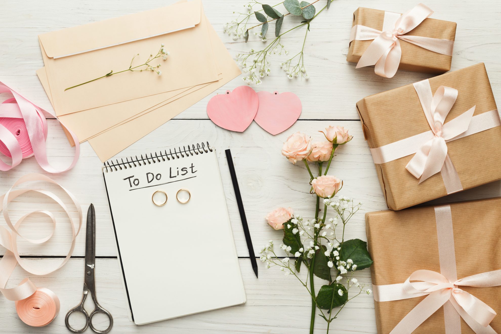 A to-do list can be seen in the image along with two wedding rings on top. Flowers surround the list along with wrapped presents. Image used in the The Top 14 Things You Need To Do Once You're Engaged article.