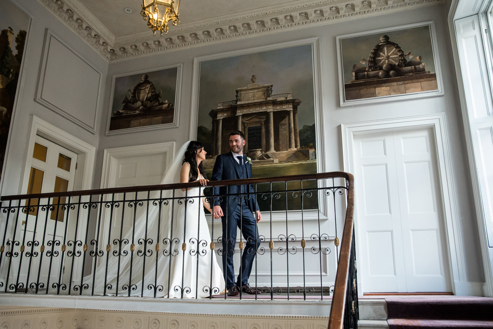 A bride and groom walking in a hallway towards the stairs. The bride is holding onto the railing. Image used in the 37 Questions To Ask Your Wedding Photographer article.